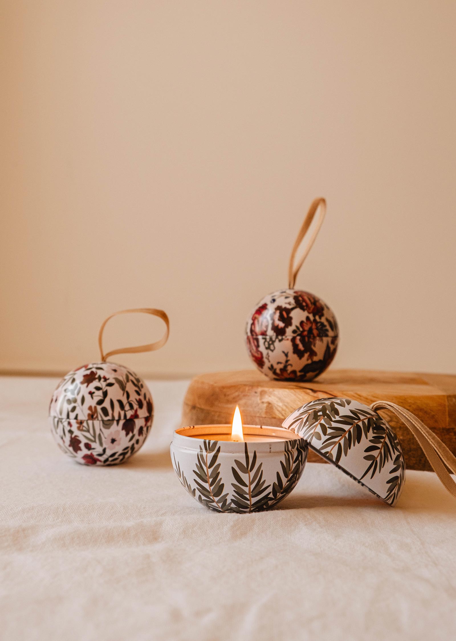 Une bougie Wintea - Christmas Ornament allumée dans un récipient à motifs jette une lueur chaleureuse, élégamment accompagnée de deux ornements assortis ressemblant à des boules de Noël. Cette charmante scène de Mimi & August repose sur une surface en tissu, créant l'ambiance parfaite pour les soirées Wintea.