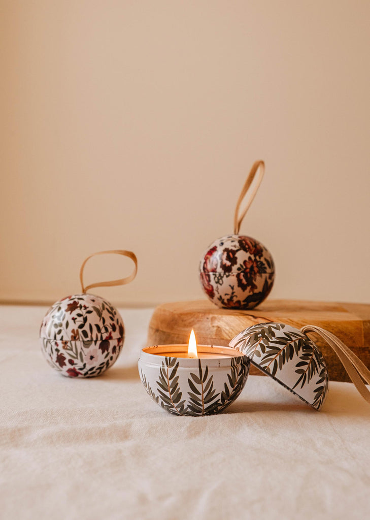 A lit Wintea - Christmas Ornament Candle in a patterned container casts a warm glow, elegantly accompanied by two matching vessel ornaments resembling Christmas baubles. This charming scene from Mimi & August rests on a fabric surface, creating the perfect ambiance for Wintea evenings.