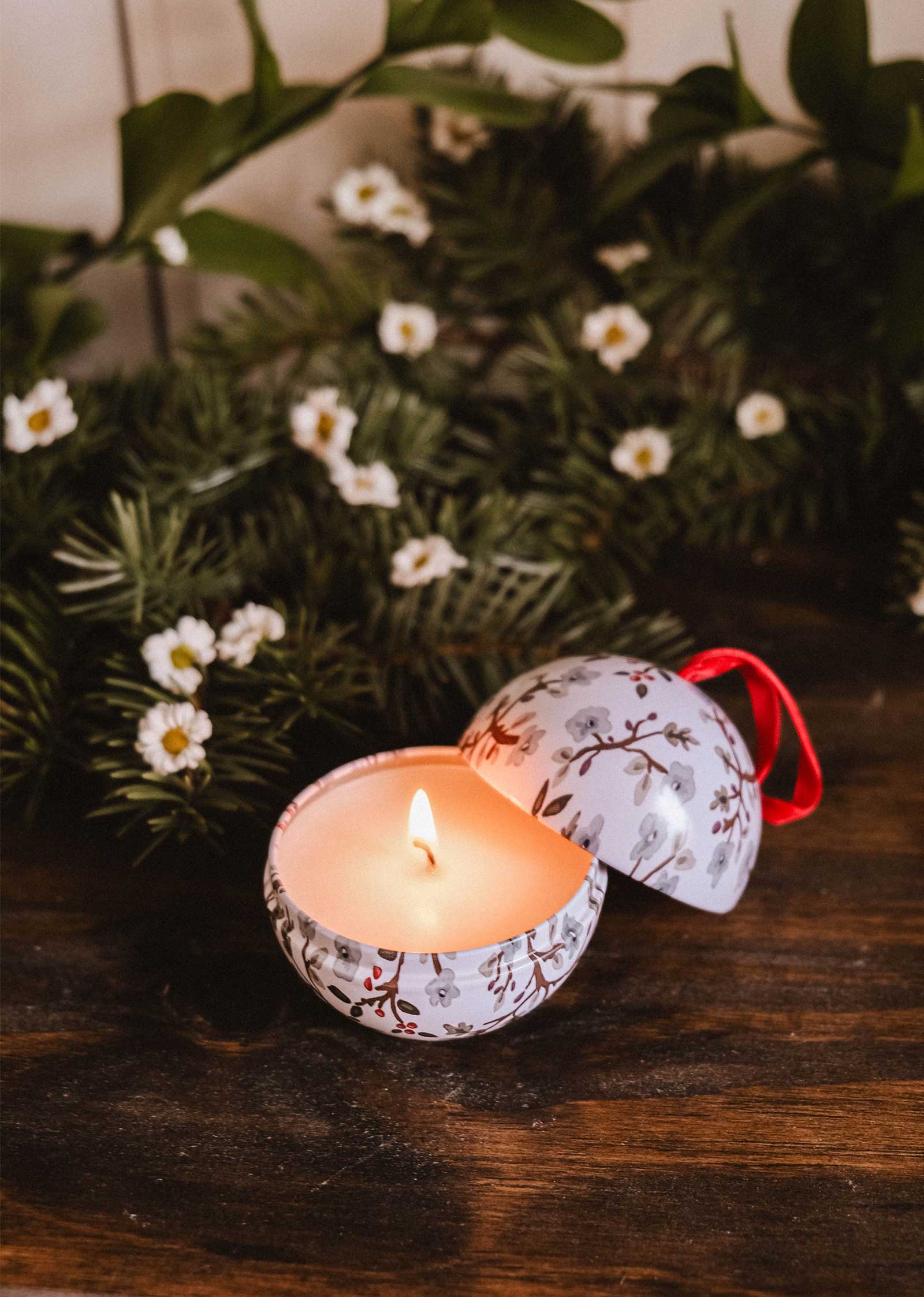 The Mimi & August Winter Flower - Christmas Ornament Candle glows in a decorative round tin on wood, surrounded by greenery and white flowers—a cozy touch for festive ambiance.