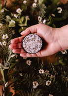 A hand holds a round tin labeled "Winter Flower - 2 oz" by Mimi & August, a floral scented candle, set against green leaves and small white flowers.