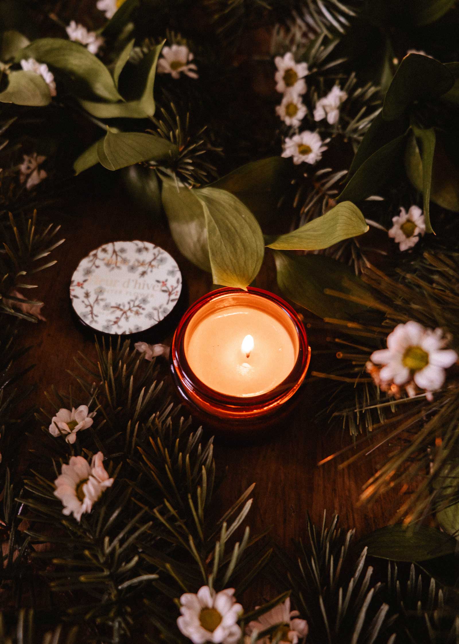 The Mimi & August Winter Flower - 2 oz candle, in a small amber jar with a floral-patterned lid, sits among green leaves and white flowers on a wooden surface.