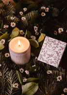 A lit Mimi & August Winter Flower - Christmas Candle in a white ribbed holder sits beside a "Fleur d’Hiver" box, surrounded by green pine branches and small white flowers.
