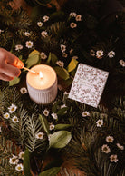 A hand lights the Mimi & August Winter Flower - Christmas Candle, scented with sandalwood and vanilla, amid pine branches, green leaves, and white flowers; a decorative "Winter Flower" box sits nearby.