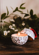 The Winter Flower - Christmas Ornament Candle by Mimi & August glows in a round, floral-patterned tin on a wooden surface with greenery and white flowers. The reusable ornament lid rests nearby.