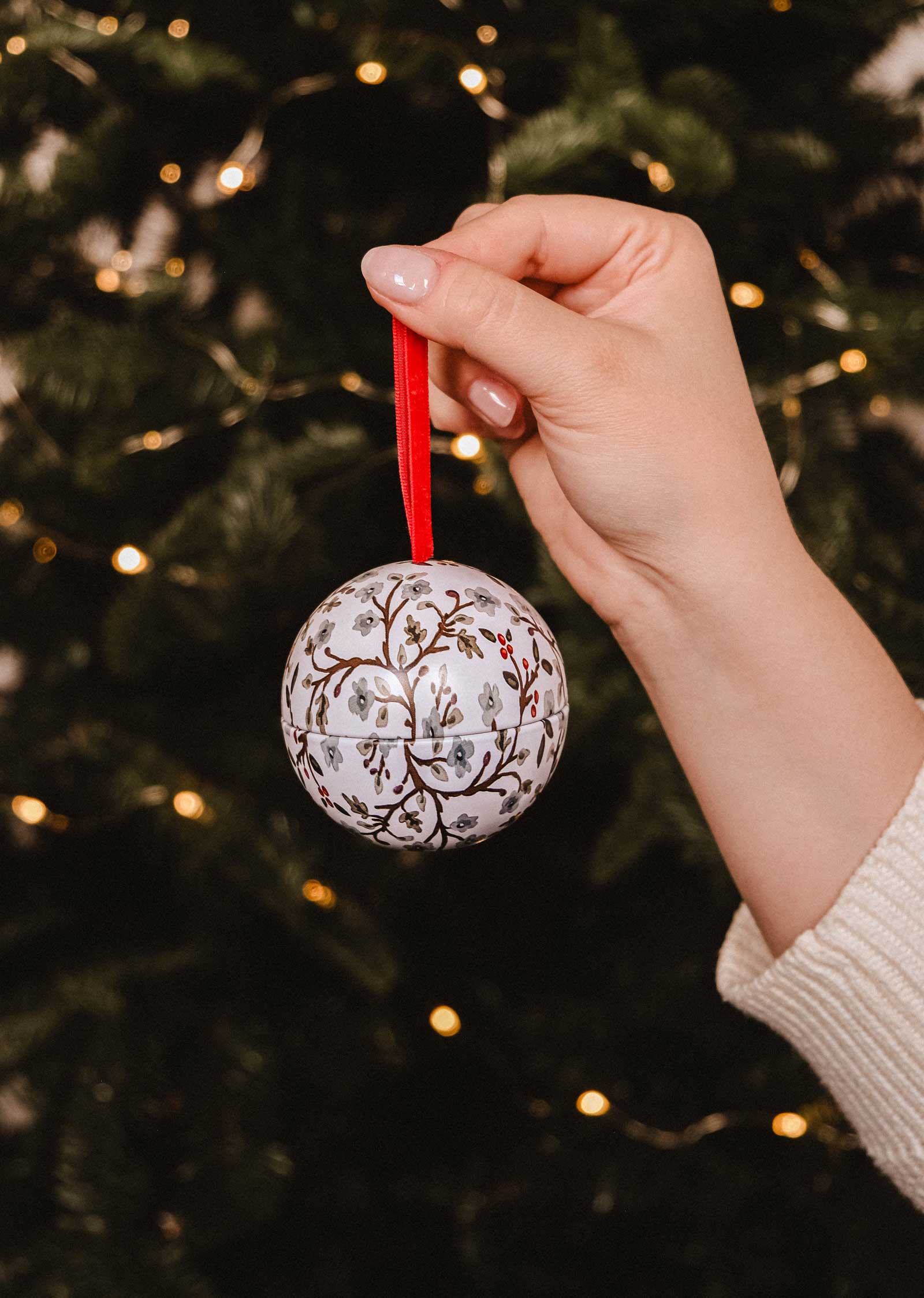 A hand holds the Mimi & August Winter Flower - Christmas Ornament Candle with floral patterns and a red ribbon, in front of a decorated Christmas tree with lights.