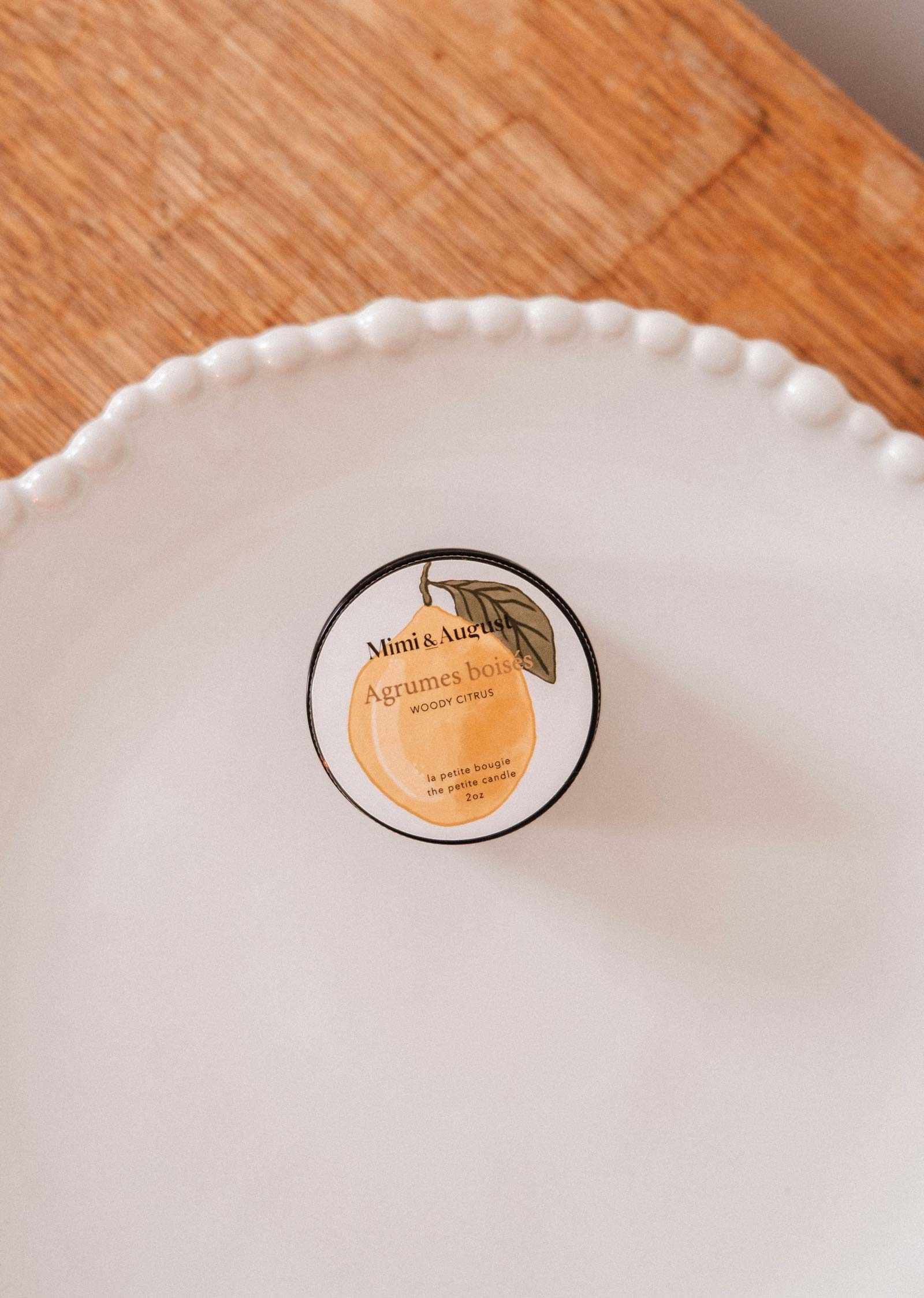 The Mimi & August Woody Citrus – 2 oz Soy Candle in a reusable glass container sits on a white, textured-rim plate atop wood, releasing lemon and cedar scents.