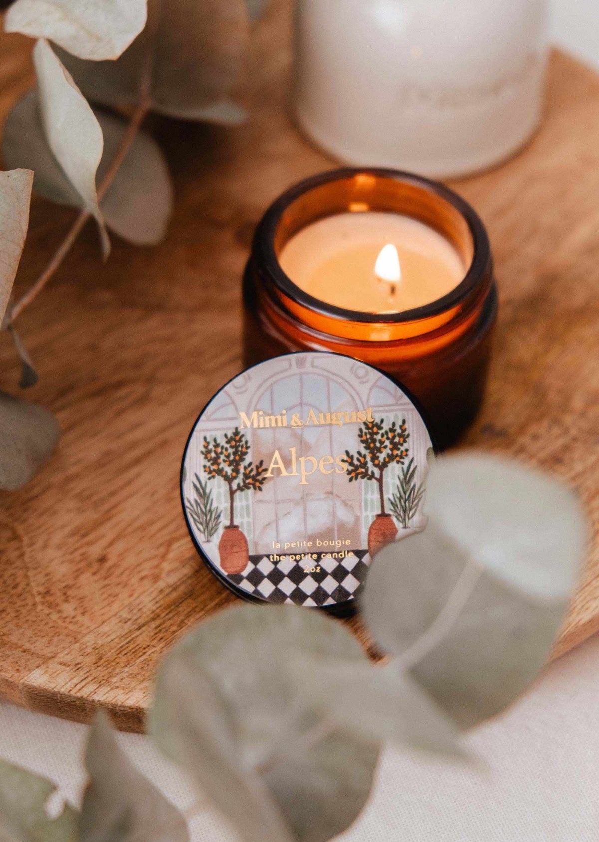 A lit amber jar candle rests on a wooden tray beside dried eucalyptus. In front, a round 2 oz tin labeled "Mimi & August Alpes" fills the room with the soothing scent and freshness of the Alps.