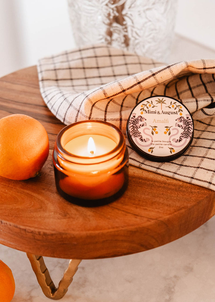 A Mimi & August Amalfi - Reusable Candle sits on a wooden board next to oranges, offering a sensory journey reminiscent of seaside holidays.