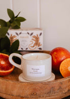 A white Mimi & August Amalfi - Reusable Candle in a cup rests on a wooden tray with blood oranges, tropical leaves, and a decorative box, capturing the seaside holiday charm.