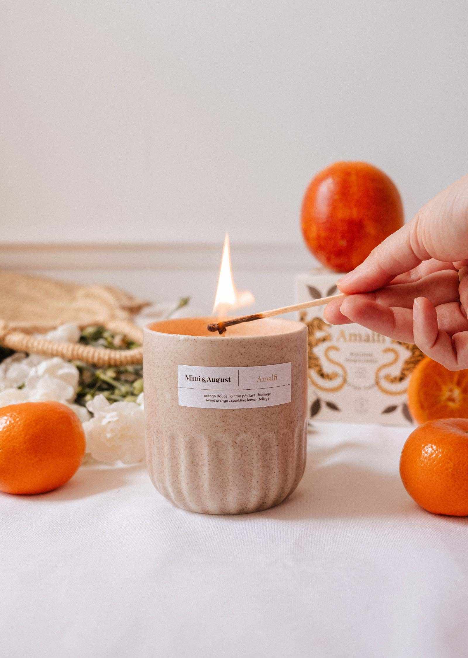 A hand lights the Amalfi - Reusable Candle by Mimi & August in a beige jar on a white surface, with oranges, a box, and tropical foliage nearby.