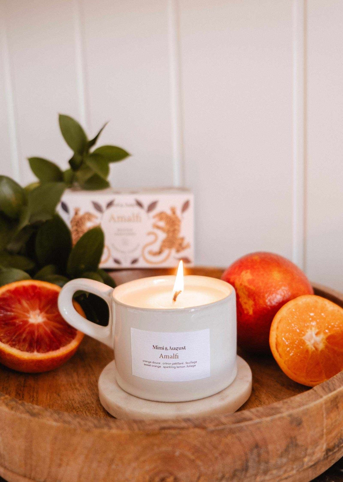 A lit Amalfi - Reusable Candle by Mimi & August, in a white cup on a wooden tray with a marble coaster, is surrounded by blood oranges and tropical foliage, evoking seaside holidays. An "Amali" box appears in the background.
