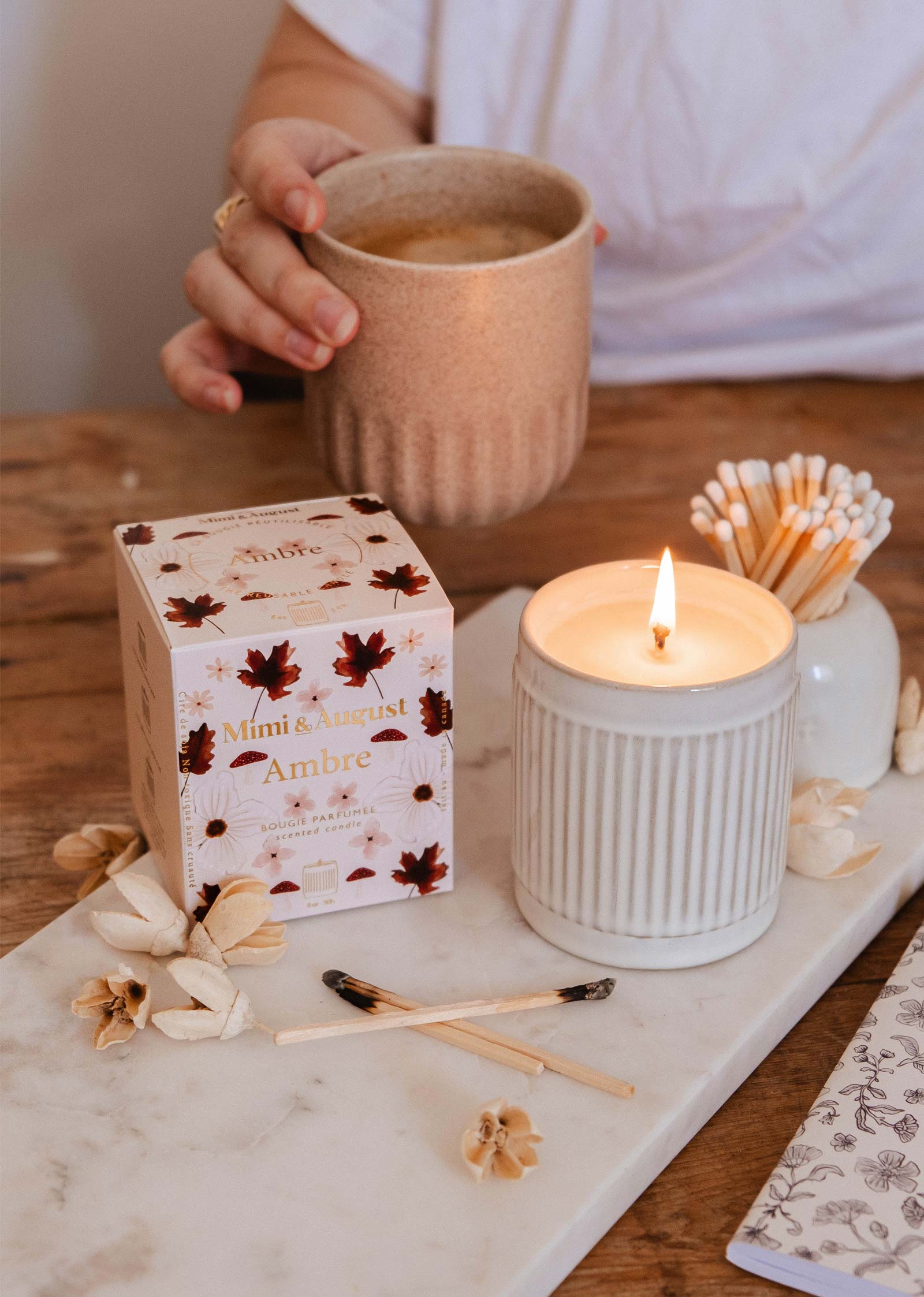 A hand holds a mug beside the Mimi & August Ambre - Reusable Candle, with scattered matches, dried flowers, and matchsticks on a marble tray—perfect for cozy fall home fragrance moments.