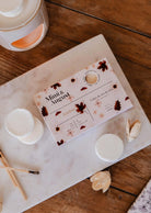 A box of Mimi & August Soy Wax Melts - Ambre rests on marble, surrounded by round wax melts, a burner, and wooden matches—ideal for filling your space with a long-lasting fragrance.