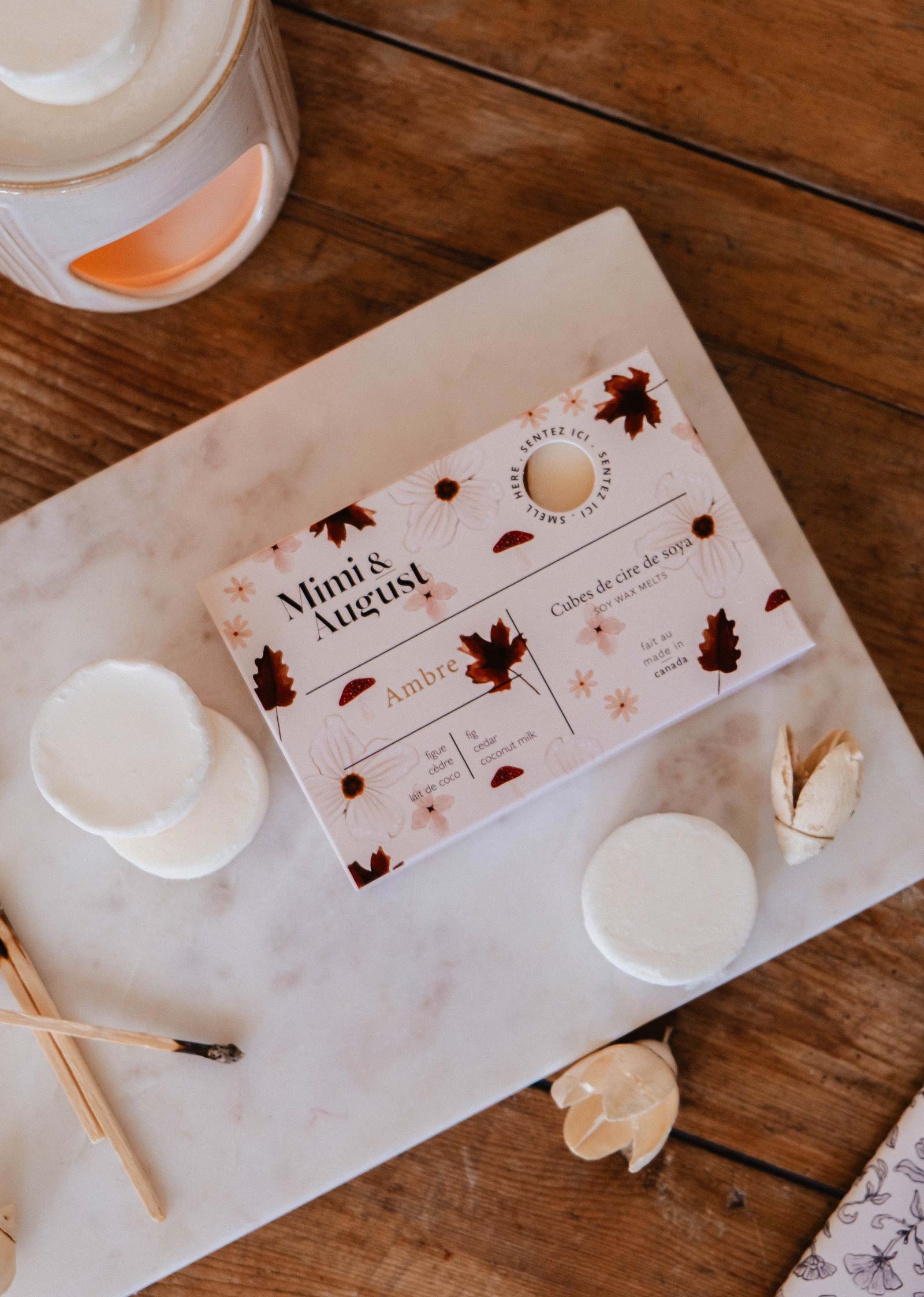 A box of Mimi & August Soy Wax Melts - Ambre rests on marble, surrounded by round wax melts, a burner, and wooden matches—ideal for filling your space with a long-lasting fragrance.