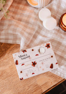 The Mimi & August Soy Wax Melts - Ambre package, featuring a fall leaf and flower design, sits on a wooden table next to a lit candle and two white soy wax melts—ideal for filling your home with cozy autumn fragrance.
