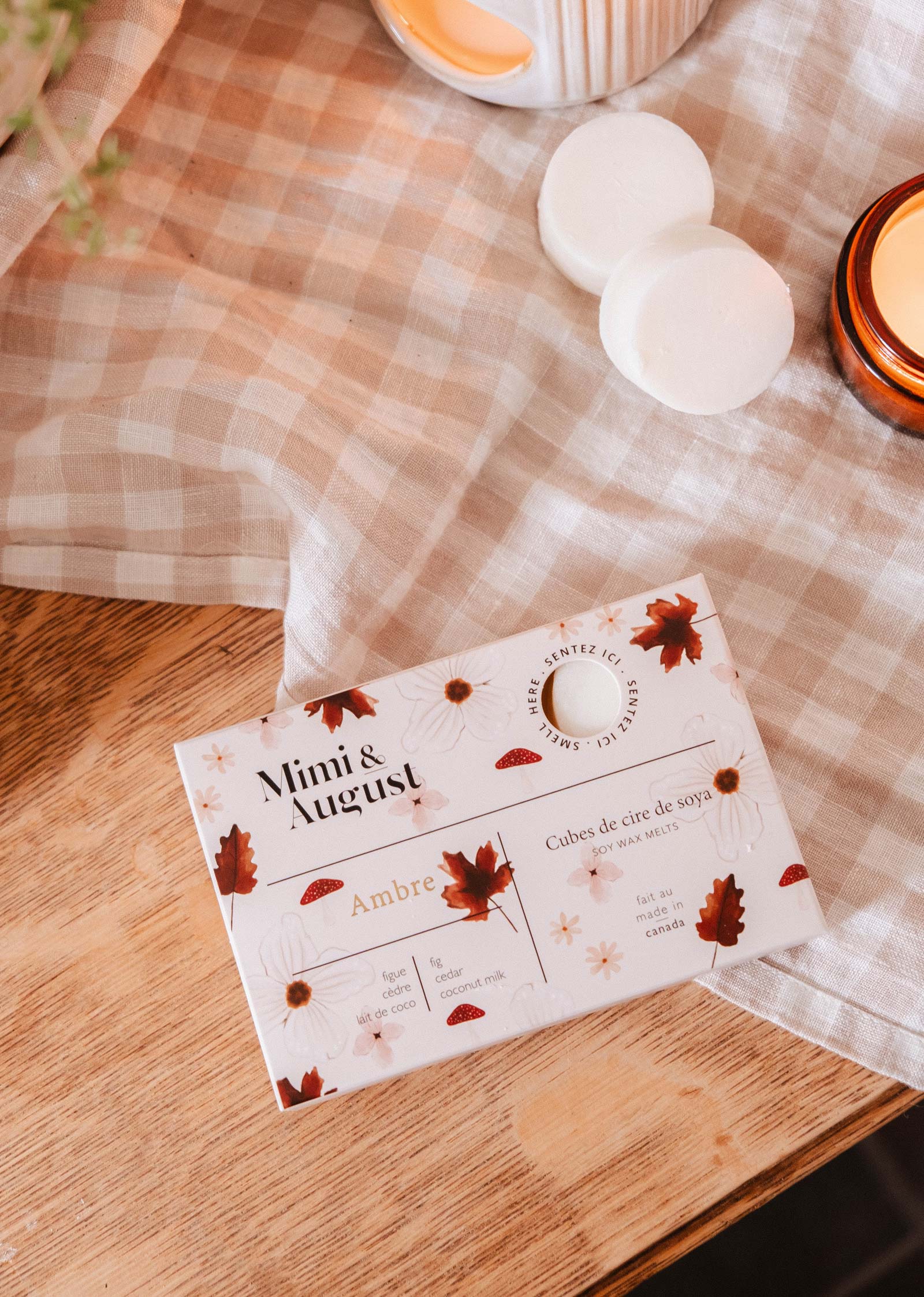 The Mimi & August Soy Wax Melts - Ambre package, featuring a fall leaf and flower design, sits on a wooden table next to a lit candle and two white soy wax melts—ideal for filling your home with cozy autumn fragrance.