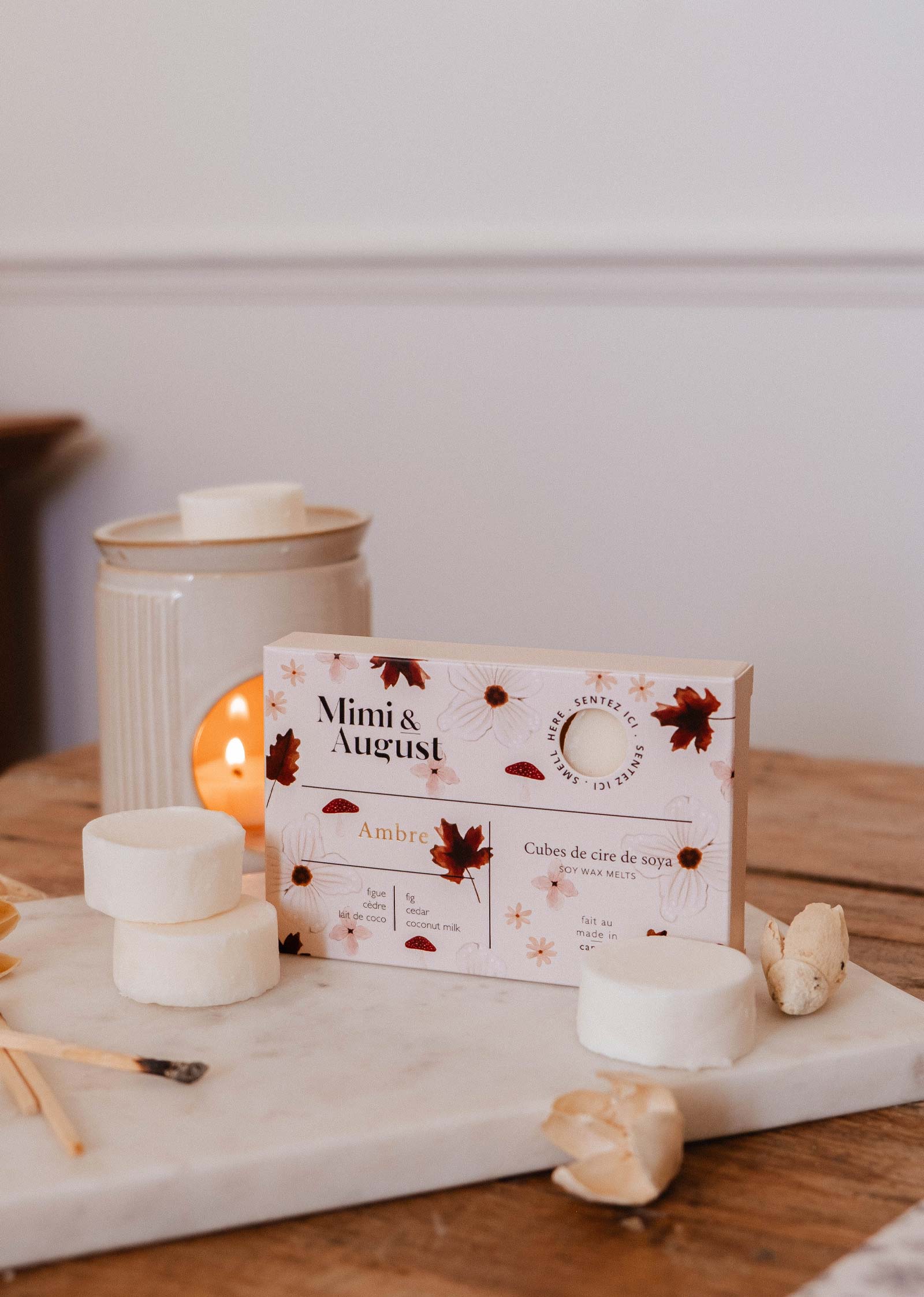 A box of Mimi & August Soy Wax Melts - Ambre sits on a wooden table, with long-lasting fragrance melts, a lit wax warmer, and a marble tray—ideal for bringing cozy autumn scent to any room.