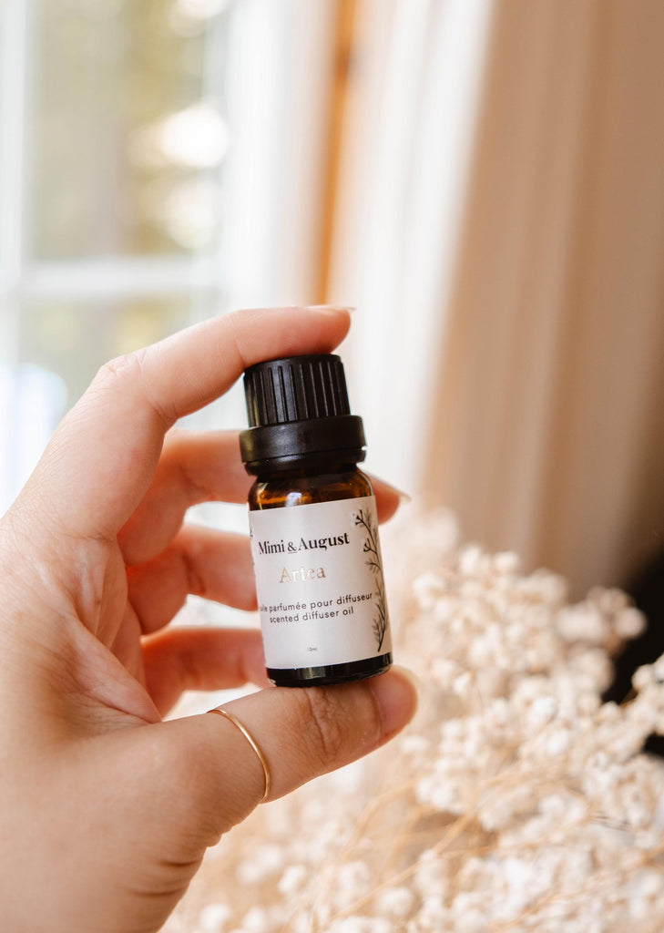 A hand holding a small bottle labeled "Mimi & August Scented Oil - Artea", against a blurred background with soft lighting and dried flowers.