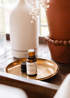 A small brown bottle labeled "Scented Oil - Artea" from Mimi & August sits on a brass plate next to a cap, emitting hints of fruity notes, with a diffuser and a terra cotta pot in the background.