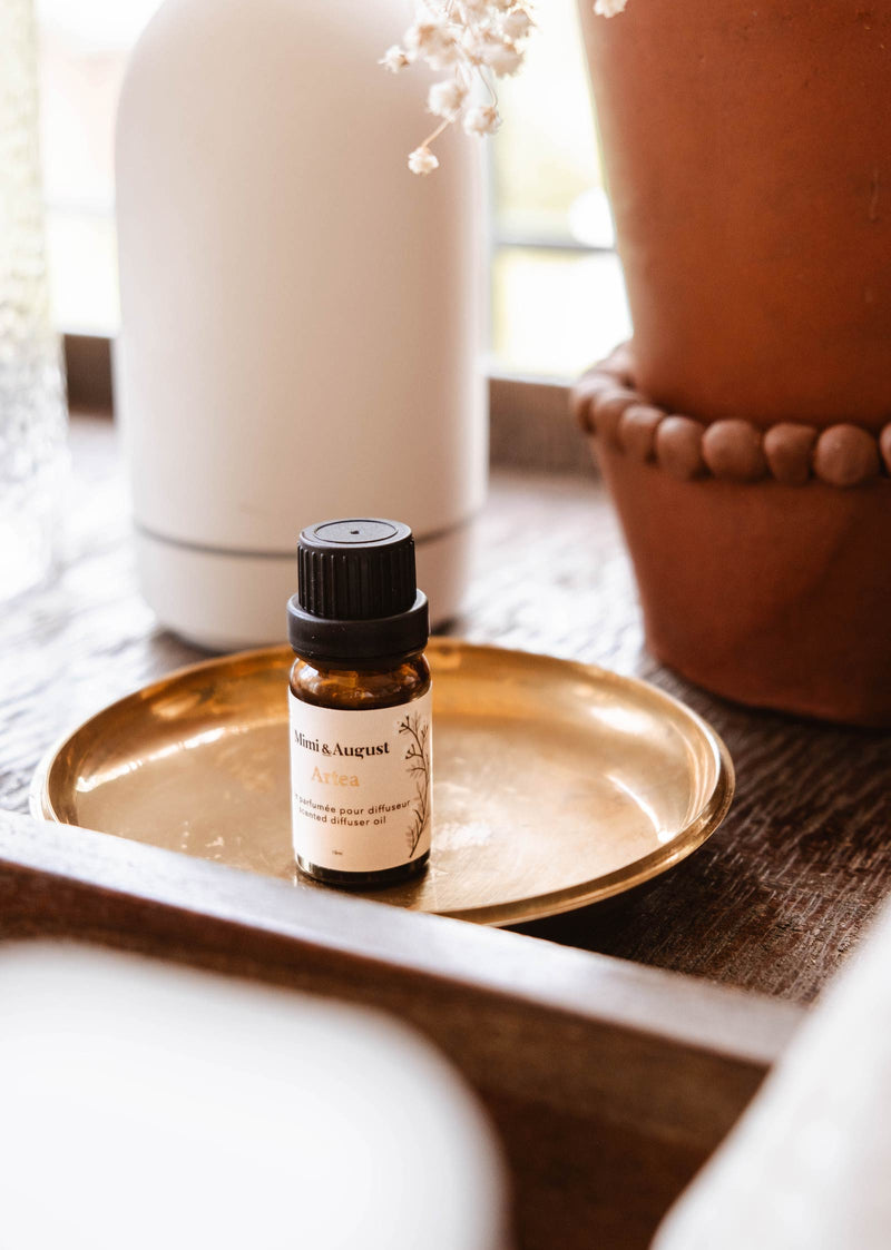 A small amber glass bottle of Scented Oil - Artea by Mimi & August with a black cap is placed on a golden plate. The bottle's label reads "Atreca" among other text. The background features blurred decor items, including a white container and a terracotta pot.