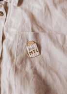 La poche d’une chemise beige met en valeur l’épinglette Bagel MTL de Mimi & August, illustrée d’un bagel et du texte « BAGEL MTL » — l’accessoire parfait pour les fans de bagels montréalais et les amoureux de bonne bouffe.