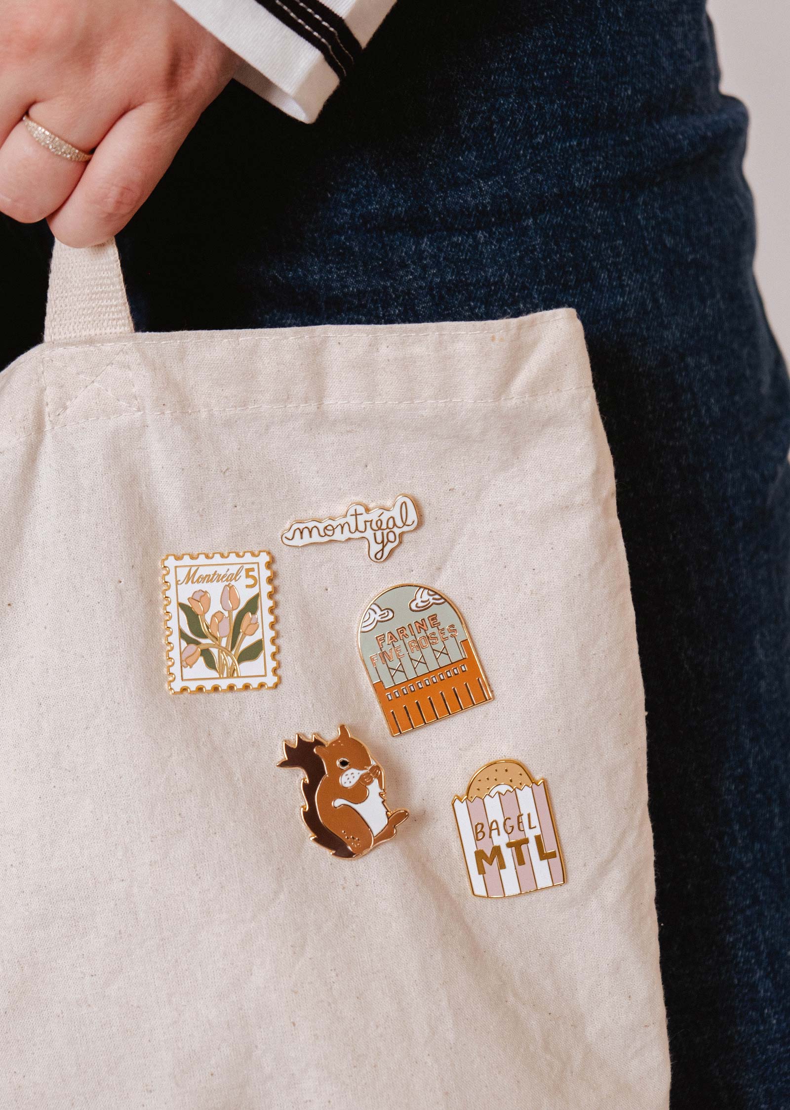 A hand holds a beige tote bag decorated with five Montreal-themed enamel pins, including the MTL Squirrel Enamel Pin by Mimi & August—featuring city buildings, flowers, a squirrel, and bagels—perfect for animal lovers.