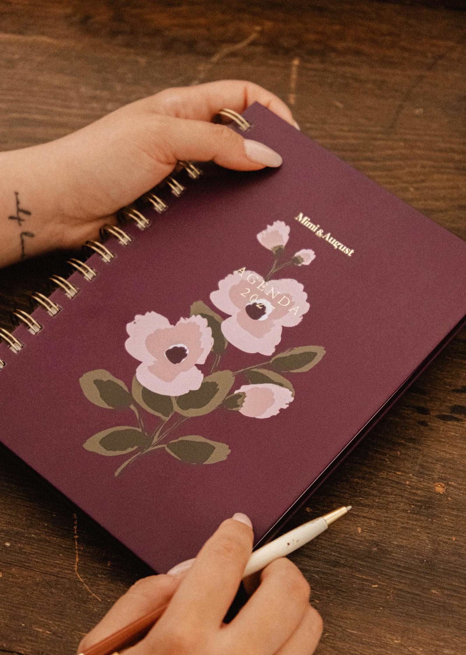 A person holds the Mimi & August Blooming Branch – 2027 Agenda, a spiral-bound planner featuring a purple floral illustration, on a wooden table, with another hand poised with a pen, ready to write.