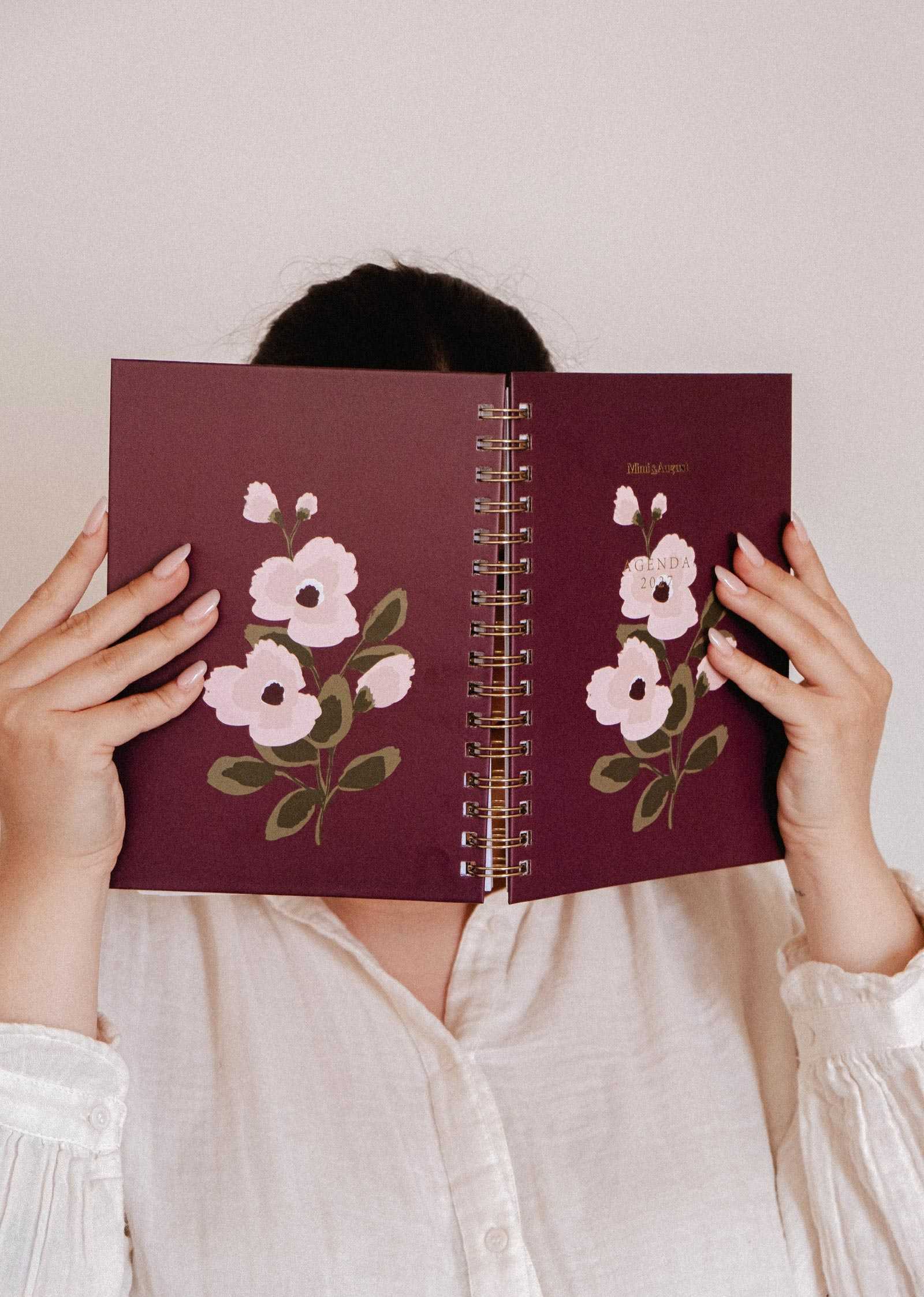 A person in a white shirt holds the Mimi & August Blooming Branch – 2027 Agenda, featuring an illustrated pink floral cover, in front of their face.