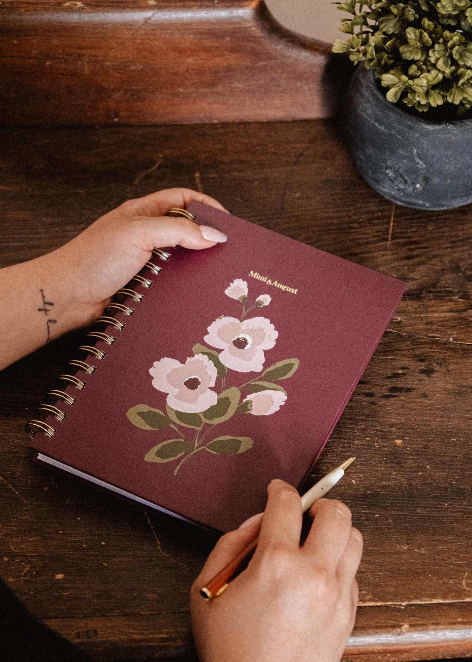 A person holds the Blooming Branch – 2027 Agenda by Mimi & August—a maroon spiral-bound planner featuring a white floral design and the brand’s name on the cover—next to a potted plant on a wooden desk.