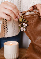 Someone adds the Mimi & August Blossom Branch Keychain to a brown leather bag, with a lit candle nearby on a wooden surface.