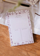The Blossom - Notepad by Mimi & August, crafted from recycled paper with sections for tasks, notes, and important items, rests on a wooden table beside a pen and a white cloth.