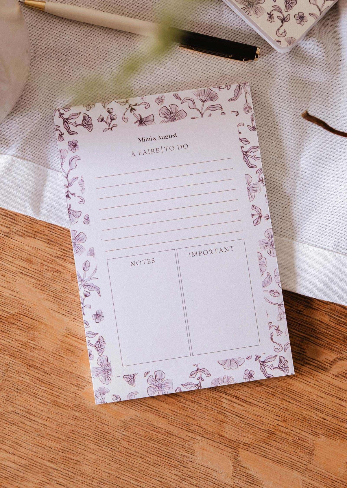 The Blossom - Notepad by Mimi & August, crafted from recycled paper with sections for tasks, notes, and important items, rests on a wooden table beside a pen and a white cloth.