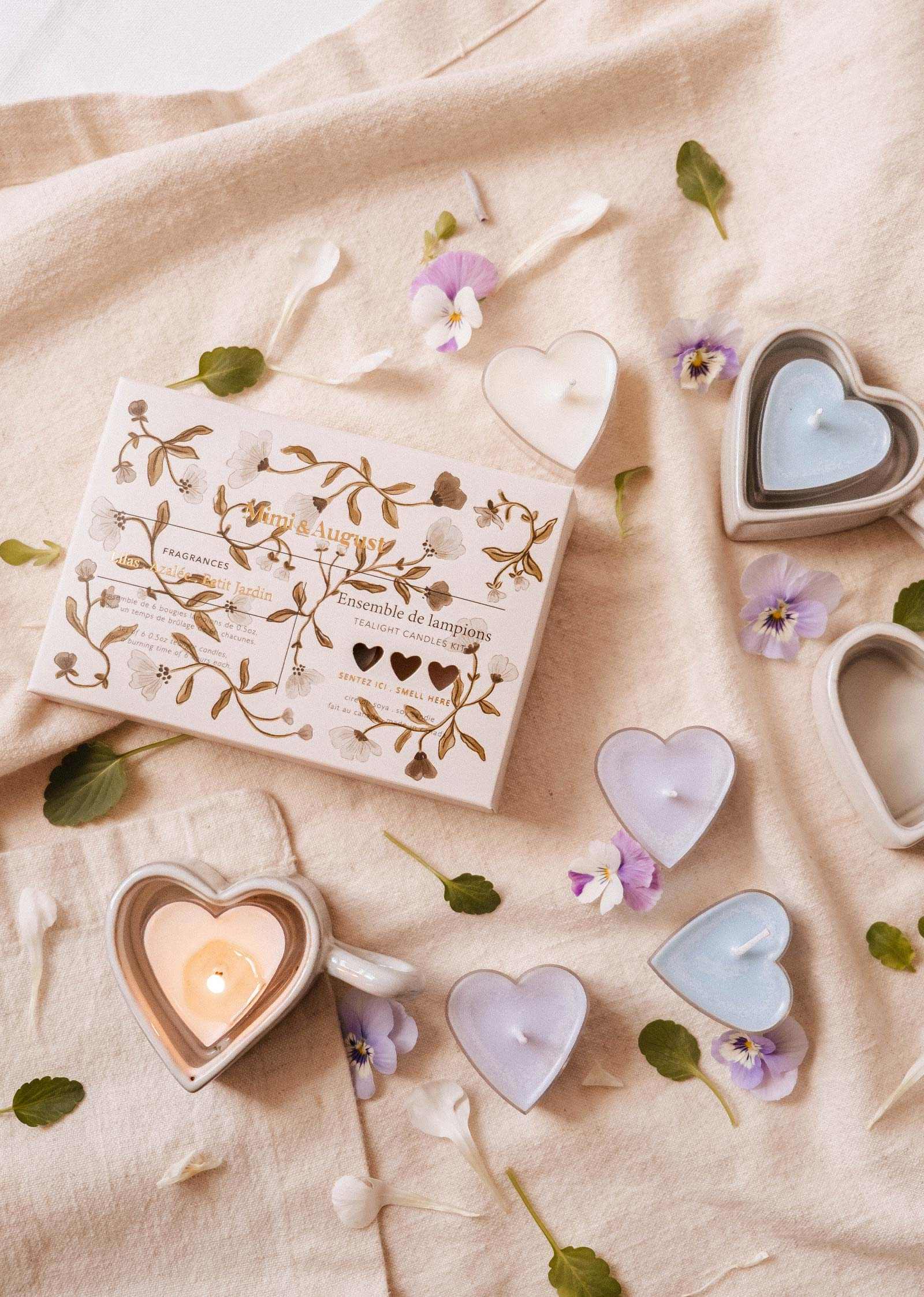 A “Blush Hearts Tealight Candle Kit” by Mimi & August rests on beige cloth, surrounded by hand-poured heart-shaped soy candles in pastel colors and scattered flower petals.