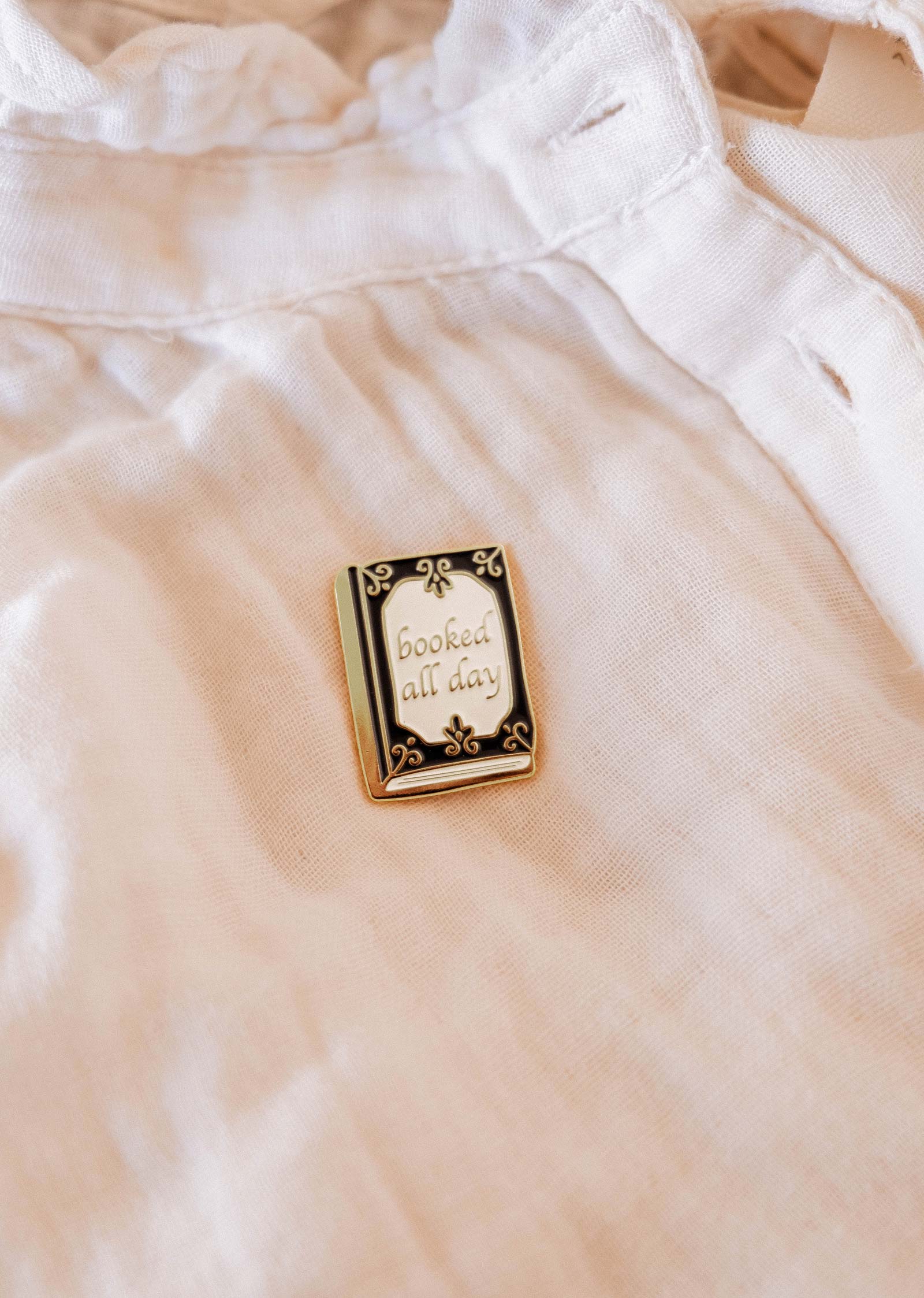 The Booked All Day Enamel Pin by Mimi & August features "booked all day" in cursive—an ideal bookworm gift and a stylish bookish accent for any white shirt or tote.