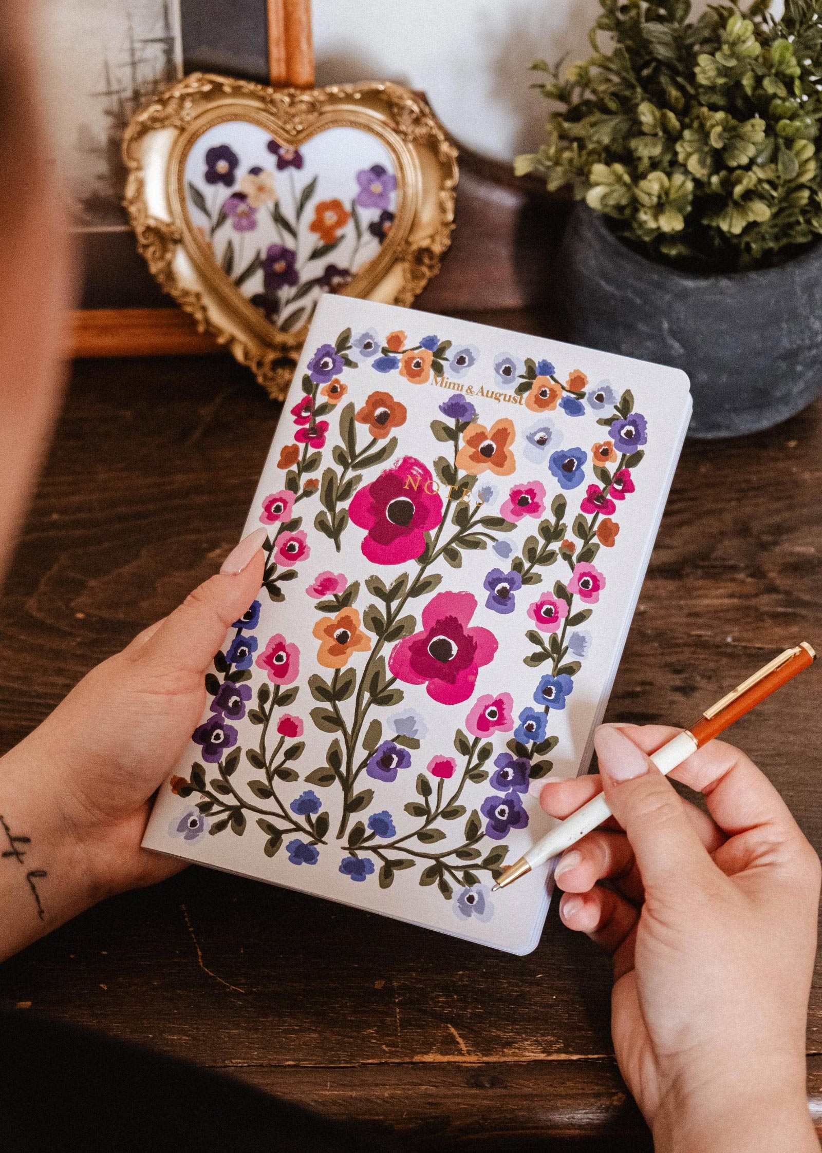 A person writes in the Bouquet floral notebook by Mimi & August, with a framed floral print and potted plant on the wooden table behind.