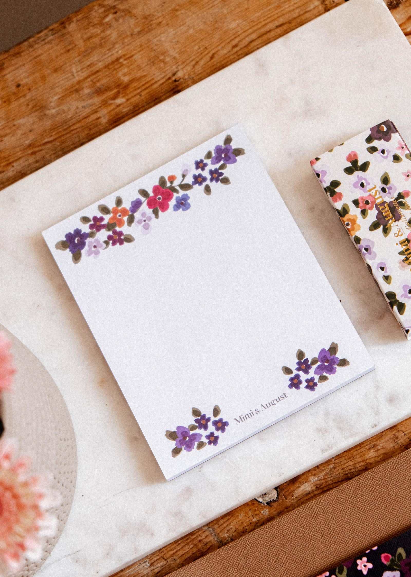 Bouquet floral notepad by Mimi & August, with hand-painted purple and pink flowers, resting on marble beside another floral item.