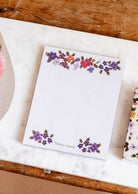 Bouquet floral notepad by Mimi & August, with hand-painted florals at the top and bottom, on a marble and wooden surface.