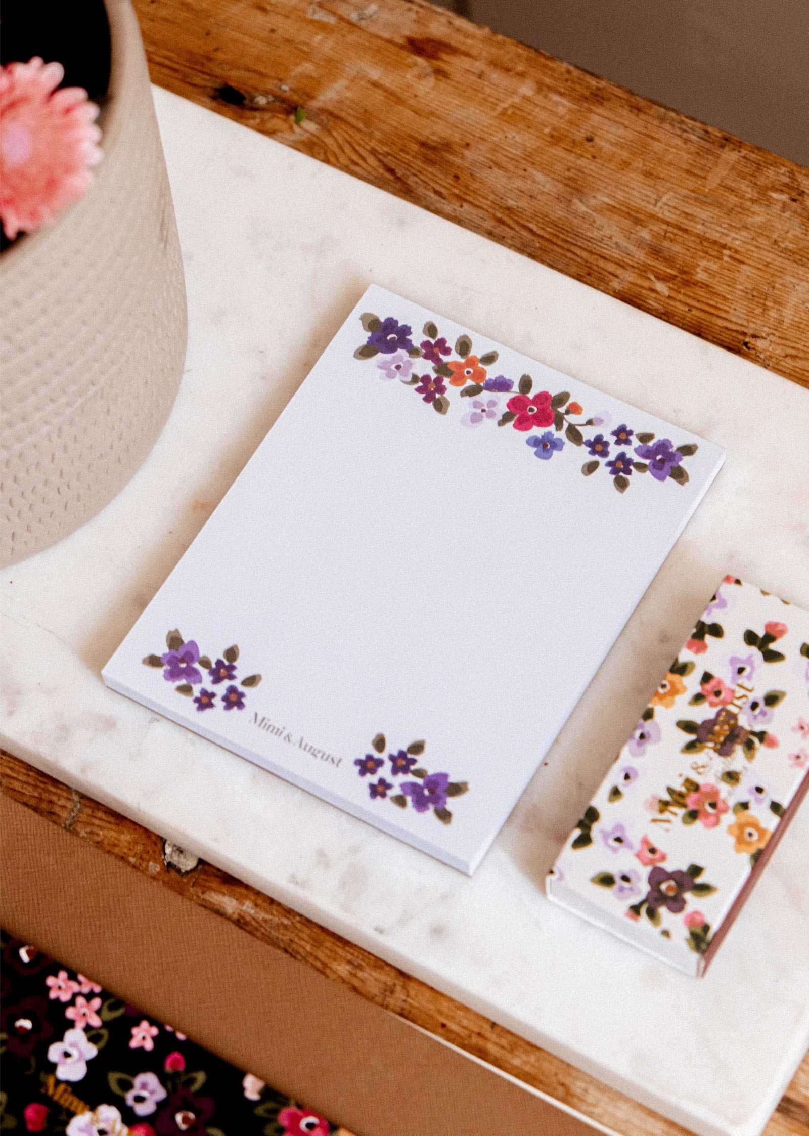 Bouquet floral notepad by Mimi & August with a matching pen on a marble surface beside a potted plant and wooden table.