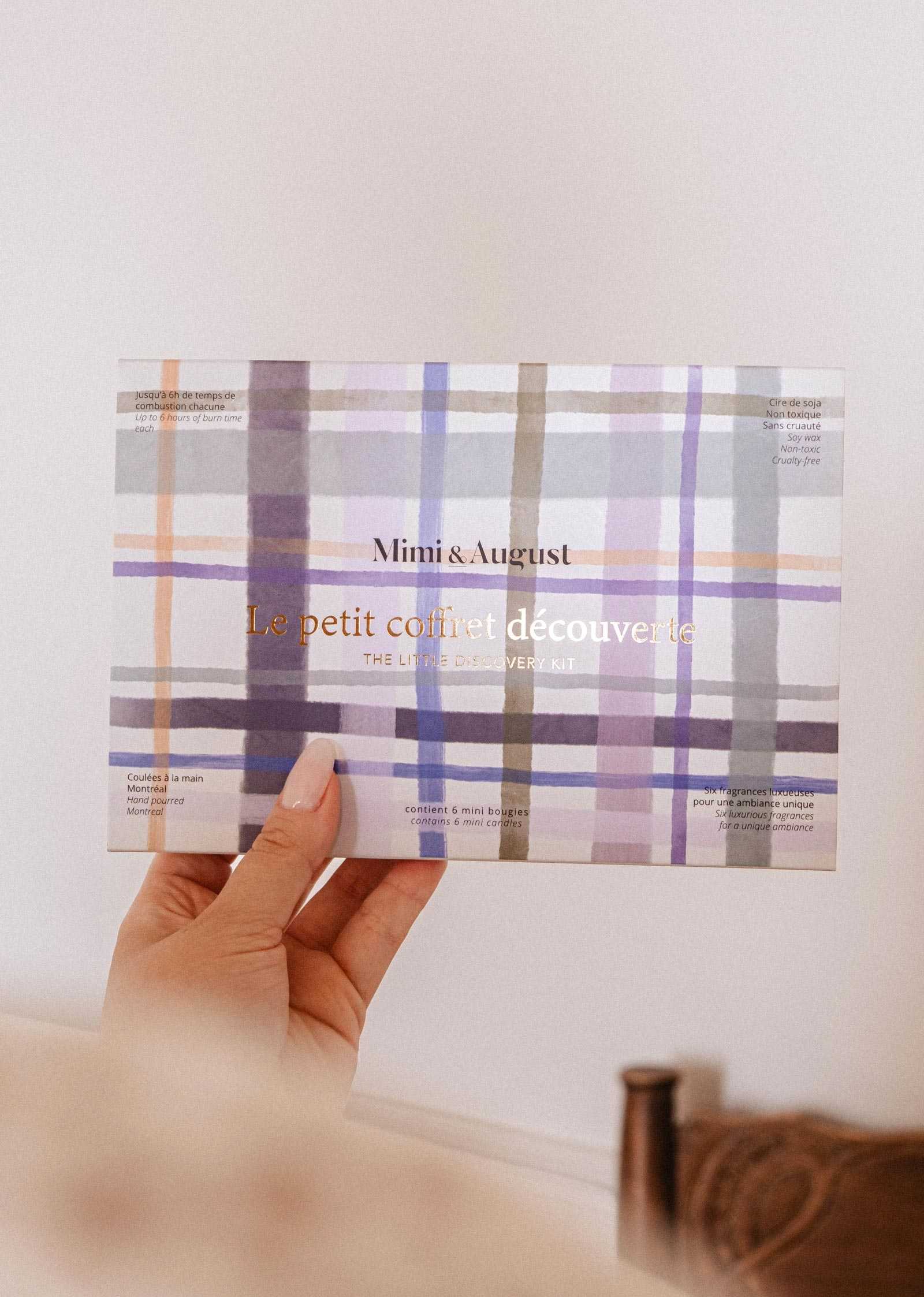 A hand holds a Mimi & August "Build Your Own Discovery Kit" box with a pastel plaid design, featuring natural soy wax candles.