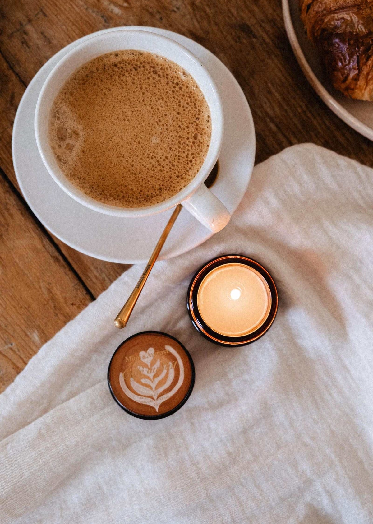 A cup of coffee and a small latte sit on a wooden table with a gold spoon, white cloth, and the Café au Lait – 2 oz Soy Candle by Mimi & August, hand-poured in Montreal for an inviting coffee-scented ambiance.