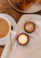 Une tasse de café, une bougie de soya Café au Lait de 2 oz de Mimi & August allumée et un croissant dans une assiette reposent sur une table en bois recouverte d'un tissu blanc.