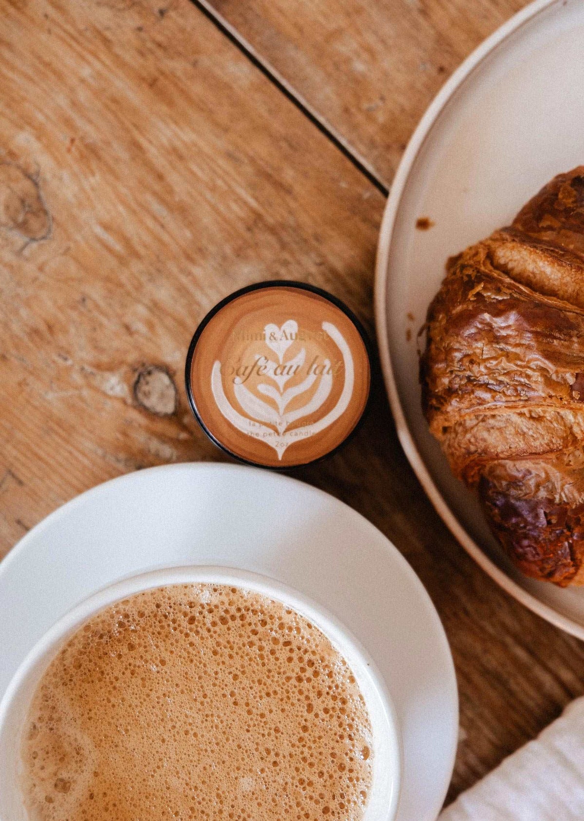 A cup of coffee with foam art, a fresh croissant, and the Mimi & August Café au lait 2 oz soy candle rest on a wooden table.