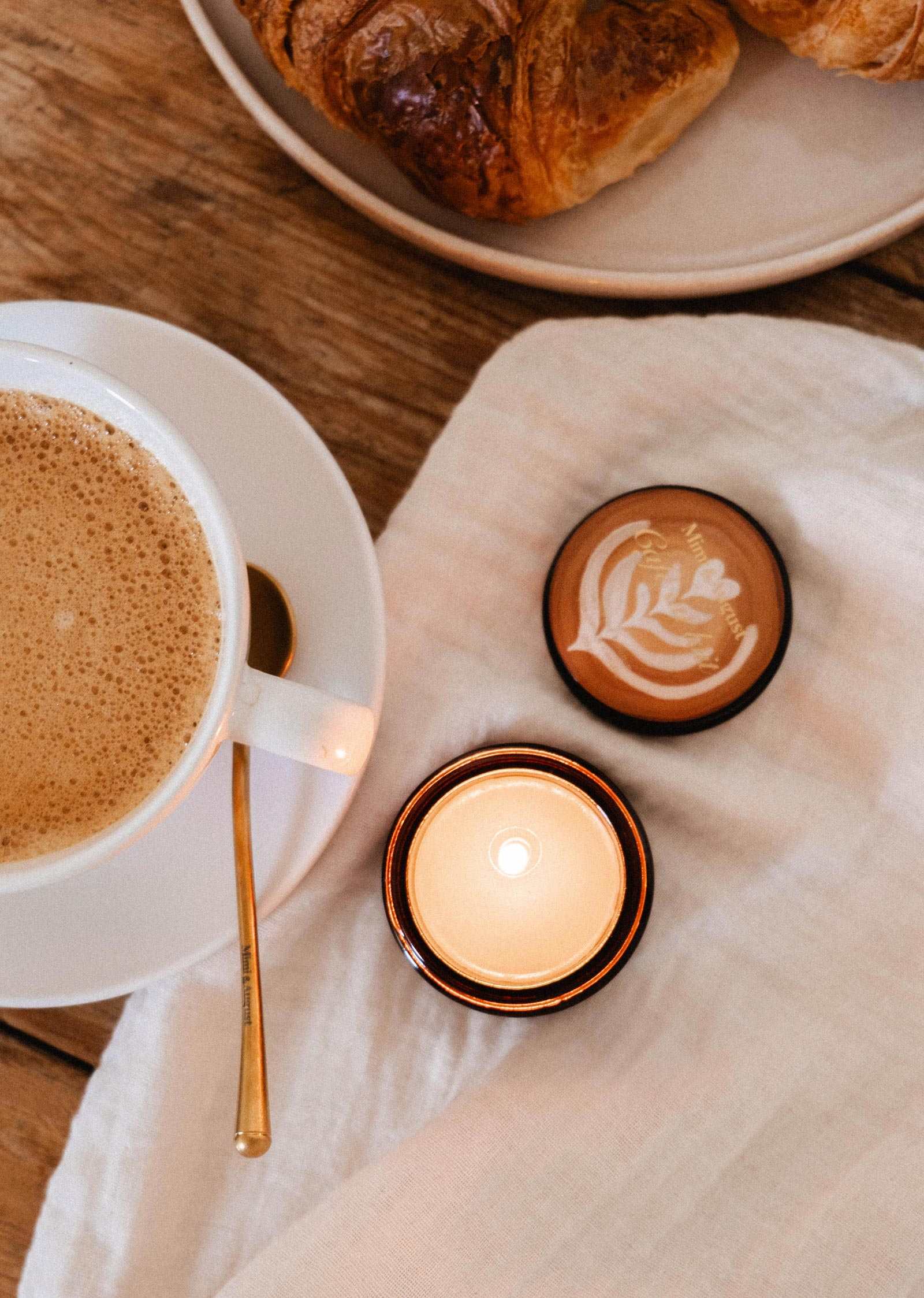 A cup of coffee, a Mimi & August Café au lait – 2 oz Soy Candle lit, and a croissant on a plate sit on a wooden table topped with a white cloth.