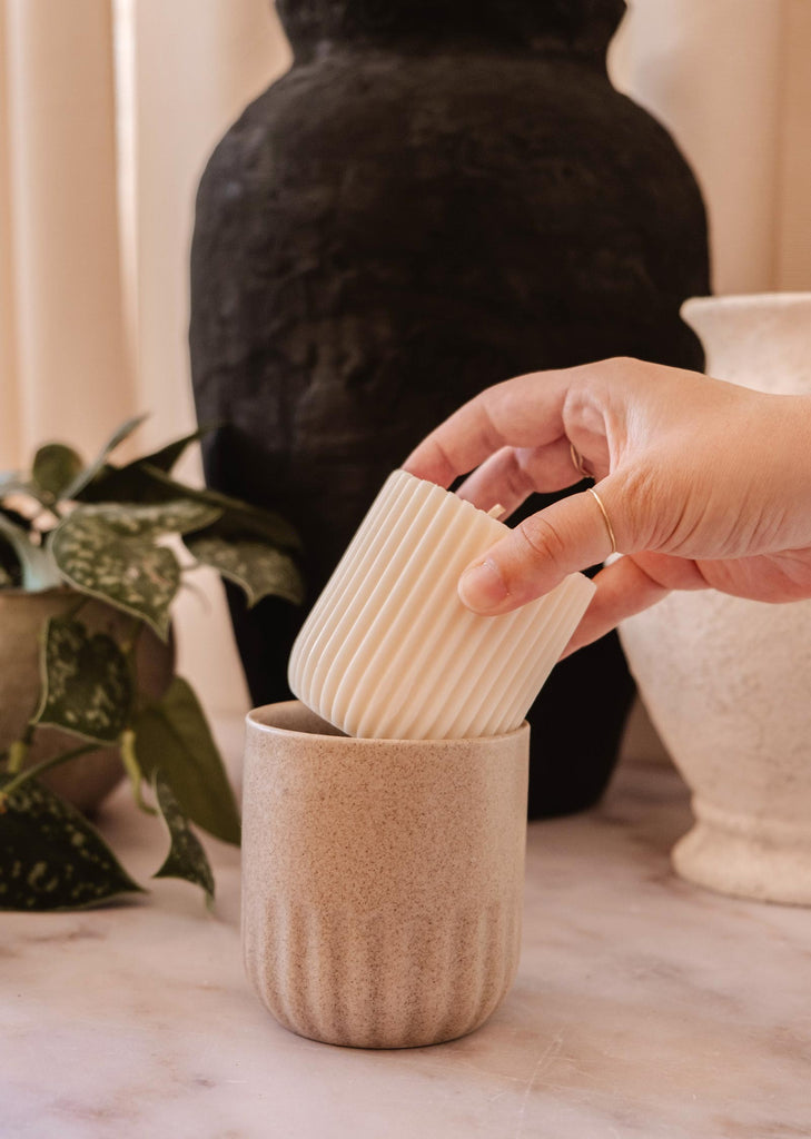 A hand is placing a white, ribbed cup inside a beige, textured cup on a marble surface, surrounded by various pottery pieces and a plant in the background. The scene evokes the serenity of sandalwood coconut vanilla, reminiscent of Candle Refill - Topaze from Mimi & August.