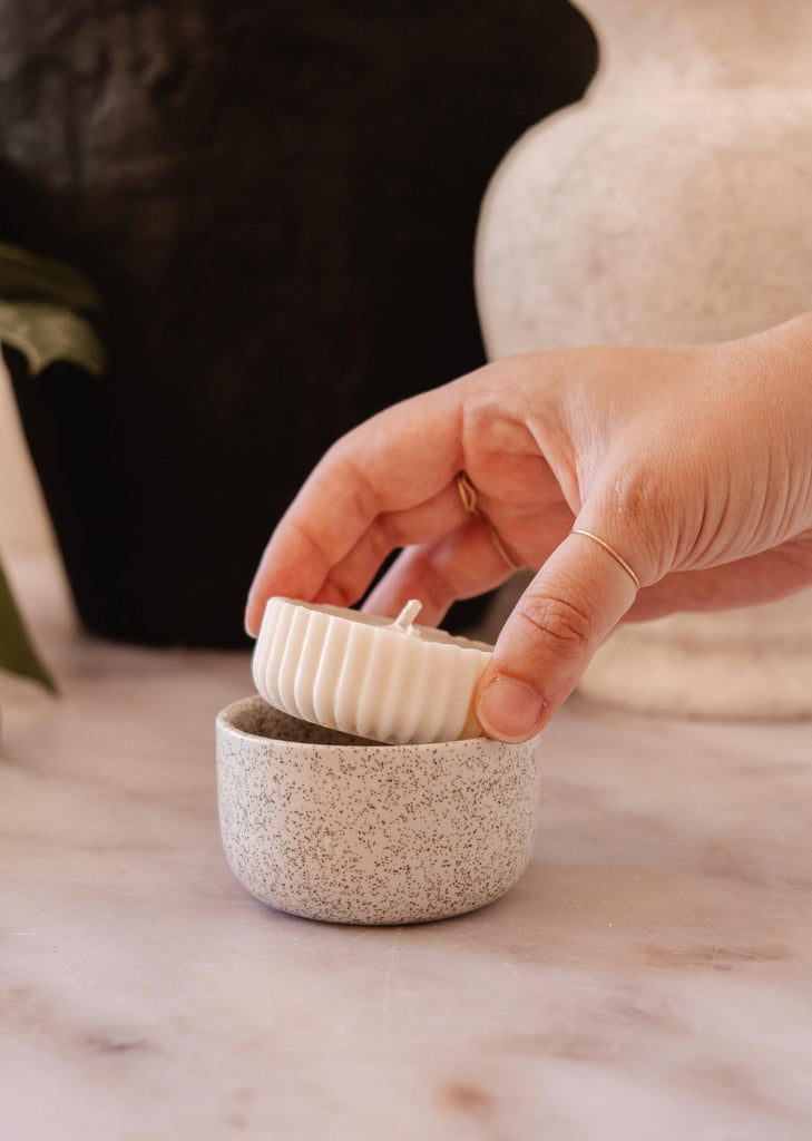 A hand is placing a Candle Refill - Petit Jardin by Mimi & August, which is white, ribbed, and round with an enchanting scent, into a speckled ceramic bowl on a marble surface.