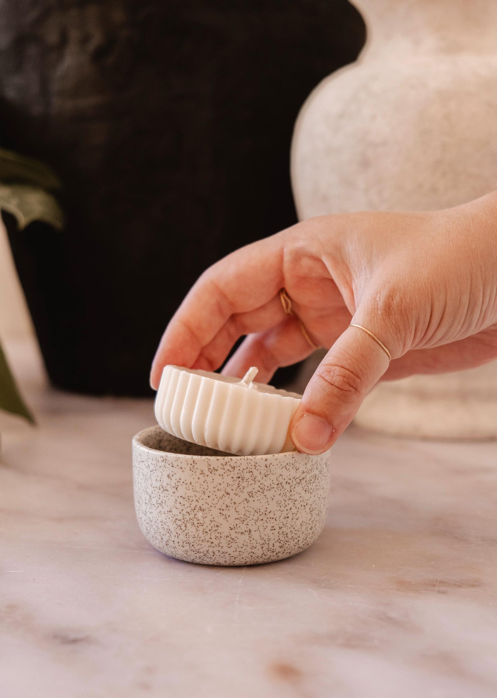 A hand placing a small, eco-friendly, white fluted Candle Refill - Topaze by Mimi & August into a speckled ceramic holder on a marble surface. The subtle scent of sandalwood coconut vanilla fills the air, creating a serene atmosphere.