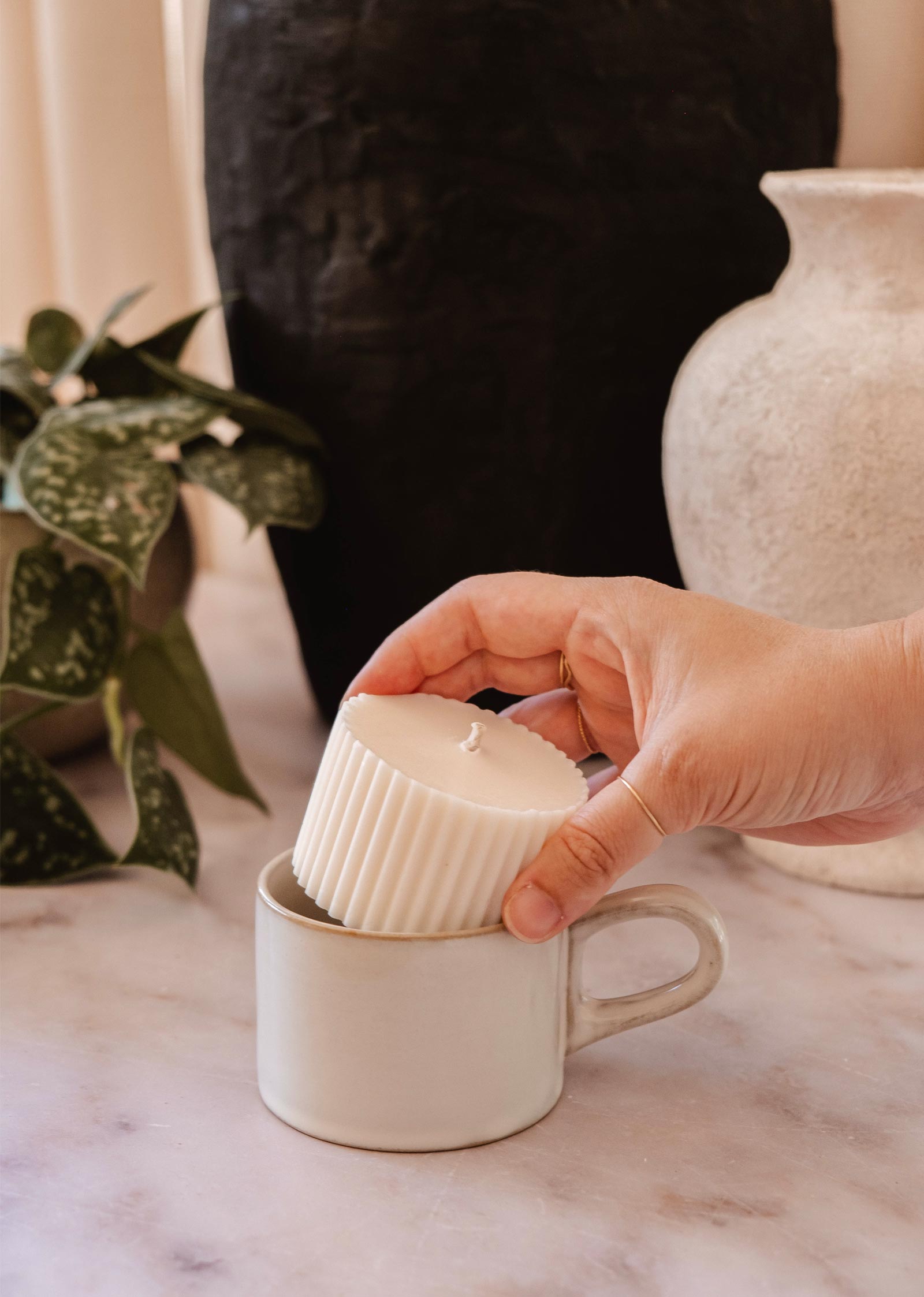 Une main place une recharge de bougie Mimi & August - Alpes à l'arôme vivifiant dans une tasse en céramique blanche sur une surface en marbre. Il y a des plantes et des pots en céramique à l'arrière-plan.