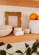 Wooden shelf with stacked ceramic bowls, a Mimi & August Candle Refill Alpes soy wax candle refill, a gold-framed picture, a white planter holding oranges, and a green plant partially visible in the foreground.