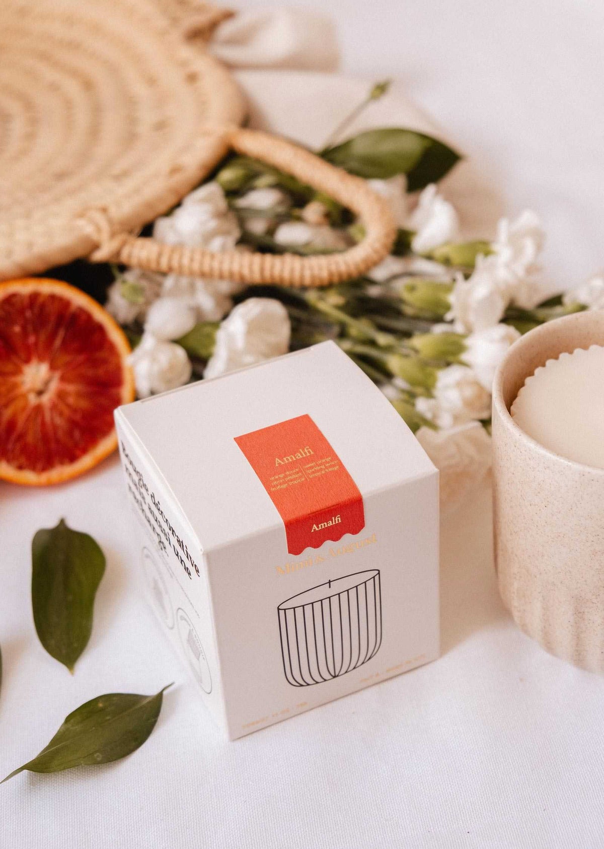 A white “Mimi & August” Candle Refill - Amalfi box sits on a white surface near a cup, sliced blood orange, green leaves, white flowers, and a woven bag—evoking Mediterranean fragrance and filled with natural soy wax.