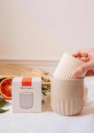 A hand places the Candle Refill - Amalfi by Mimi & August, a ribbed white natural soy wax candle, into a beige ceramic holder beside its box, with blood orange slices and greenery in the background.